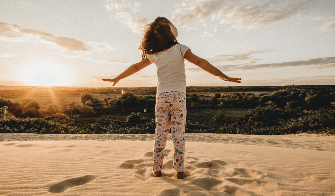 Ein kleines Mädchen steht mit ausgebreiteten Armen am Strand und sieht in den Himmel
