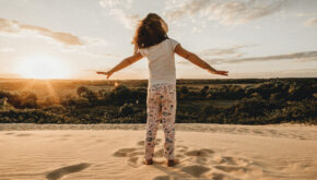 Ein kleines Mädchen steht mit ausgebreiteten Armen am Strand und sieht in den Himmel