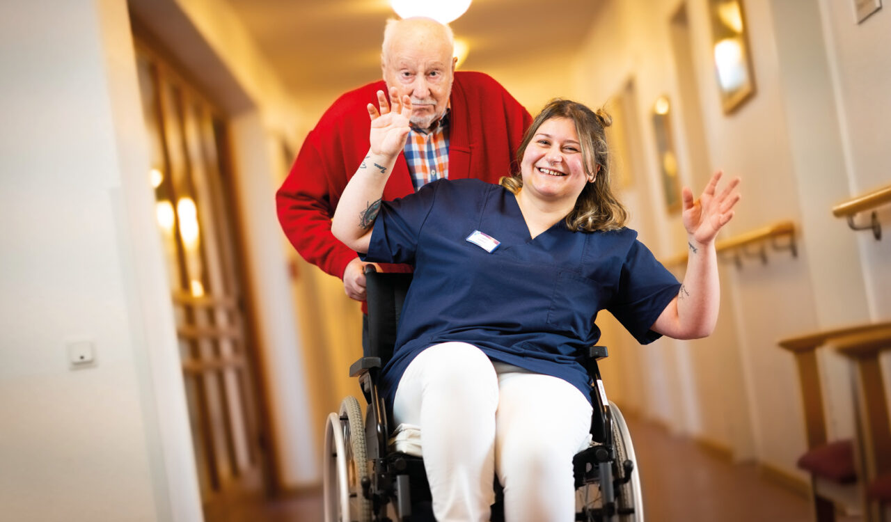 Ein Heimbewohner fährt eine lachende Pflegerin im Rollstuhl durch ein Pflegeheim