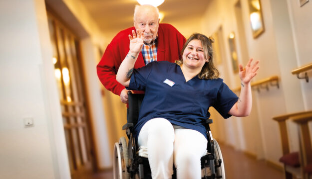 Ein Heimbewohner fährt eine lachende Pflegerin im Rollstuhl durch ein Pflegeheim